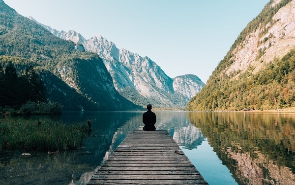 a person sits at the end of a dock looking out onto a peaceful lake surrounded by mountains