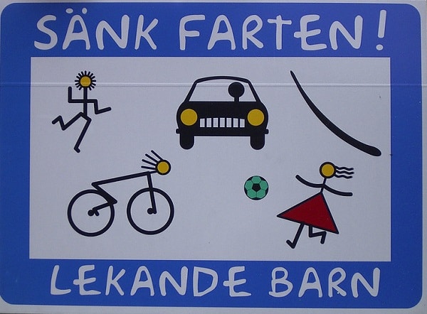 a swedish-language road sign says "sank farten; lekande barn" with drawings of kids playing and cyclists in the road